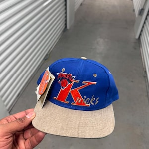 Puede incluir: Una gorra de béisbol azul y beige con el logotipo de los Knicks de Nueva York. La gorra tiene una corona azul, una visera beige y el logotipo del equipo en rojo y naranja. La palabra "Knicks" está bordada en blanco.