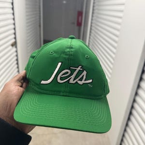 Może przedstawiać: Zielona czapka bejsbolowa z haftowanym białym tekstem "Jets" z symbolem znaku towarowego.