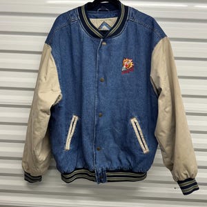 May include: A blue denim bomber jacket with tan sleeves. The jacket has a button-up front, two front pockets, and a ribbed hem and cuffs. A logo is embroidered on the chest. The jacket is hanging on a black hanger.