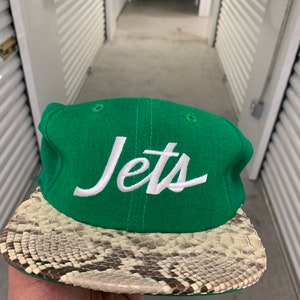 Pode incluir: Um boné de beisebol verde com um logotipo "Jets" branco e uma aba com padrão de pele de cobra.