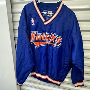 May include: A blue long-sleeve pullover jacket featuring the New York Knicks logo in orange and white lettering across the chest. The garment has a V-neck collar with white and orange stripes, matching striped cuffs, and an NBA logo on the upper right.