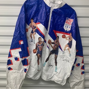 Puede incluir: Chaqueta cortavientos blanca y azul con un logotipo olímpico de EE. UU. rojo, blanco y azul. La chaqueta presenta imágenes de jugadores de baloncesto del Dream Team de 1992, incluidas las firmas de Charles Barkley, Larry Johnson y Scottie Pippen.
