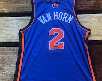 Puede incluir: Una camiseta de baloncesto azul con ribetes naranja y negro. La parte posterior de la camiseta tiene el nombre "VAN HORN" y el número "2" en naranja. La camiseta está firmada con tinta negra.