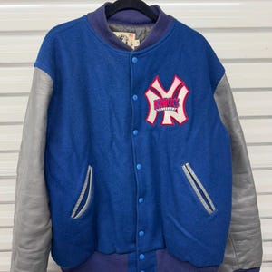 May include: Blue and gray varsity jacket with a button-up front. The jacket features a white and red logo on the chest and gray leather sleeves. The collar, cuffs, and waistband are a darker blue.