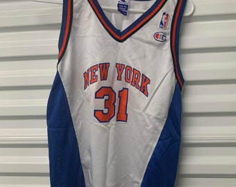 May include: White basketball jersey with blue and orange accents. The jersey has "NEW YORK" and the number "31" on the front. The jersey is made by Champion and has the NBA logo on the front.