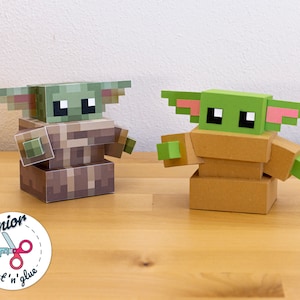 Könnte beinhalten: Zwei Papierfiguren eines grünen und braunen Charakters mit großen Ohren. Die Figuren sind aus Pappe und werden mit Laschen und Schlitzen zusammengebaut. Die Figuren stehen auf einer Holzoberfläche. Der Text "Junior Cut 'n' glue" ist in der linken unteren Ecke des Bildes sichtbar.