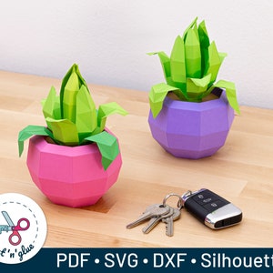 May include: Two papercraft succulent plants in pots. The pots are geometric and have a pink and purple colour scheme. The plants are green and have a 3D look. The text "PDF. SVG. DXF. Silhouette" is at the bottom of the image.