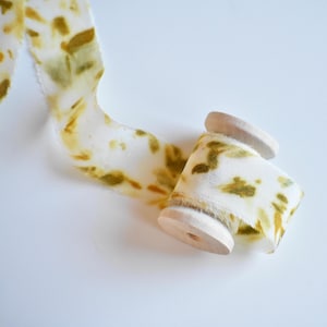 Puede incluir: Una cinta blanca con manchas verde oliva y amarillas enrollada parcialmente en un carrete de madera. La cinta tiene un aspecto suave y texturizado, y el carrete es de madera clara.