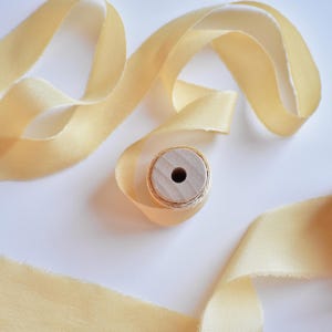 Puede incluir: Una bobina de cinta de satén amarillo pálido con un centro de madera. La cinta está dispuesta en un patrón decorativo sobre una superficie blanca.
