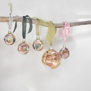 May include: A collection of glass Christmas ornaments with abstract designs in shades of gold, brown, and pink. Each ornament hangs from a natural branch with a ribbon in various colors, including green, pink, and cream.