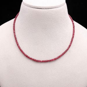 Op de afbeelding: Een delicate ketting bestaande uit kleine, ronde, dieprode kralen. De ketting wordt gepresenteerd op een witte mannequin buste tegen een donkere achtergrond. De kralen lijken gelijkmatig van grootte en dicht op elkaar geregen.