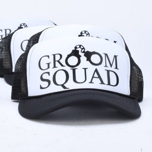 May include: Four black and white trucker hats with a white mesh back. The front of the hats have a black graphic of a soccer ball and the text "Groom Squad".