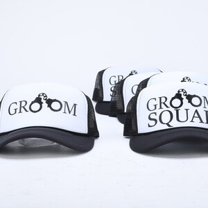 May include: Five black and white trucker hats with mesh back panels. The hats have a white front panel with black text and a graphic of handcuffs. The text on the hats reads "Groom" and "Groom Squad".