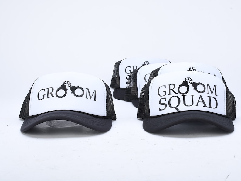 May include: Five black and white trucker hats with mesh back panels. The hats have a white front panel with black text and a graphic of handcuffs. The text on the hats reads "Groom" and "Groom Squad".