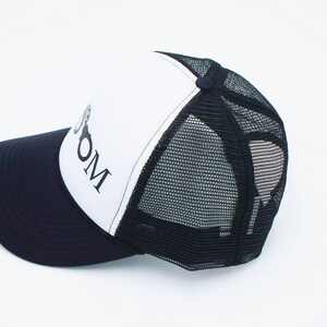 May include: A white and black trucker hat with a black mesh back. The front of the hat has a black and white logo that says "BOOM".