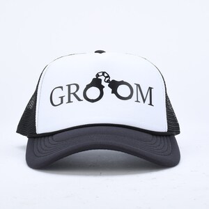 May include: A white and black trucker hat with the word "GROOM" printed on the front in black lettering. The word "GROOM" is centered on the hat and is surrounded by a black silhouette of handcuffs.