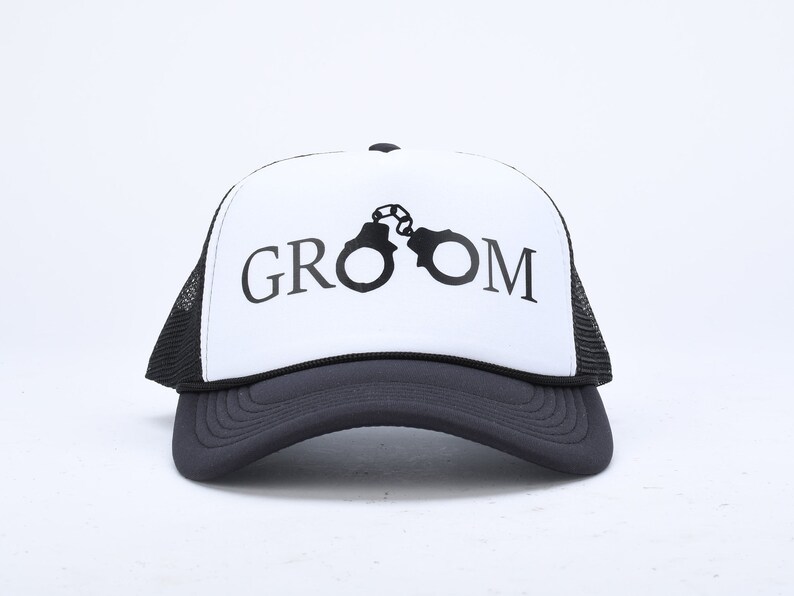 May include: A white and black trucker hat with the word "GROOM" printed on the front in black lettering. The word "GROOM" is centered on the hat and is surrounded by a black silhouette of handcuffs.