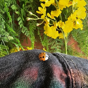 May include: A close-up of a black denim jacket with a small ladybug pin on the front. The ladybug is orange with black spots and white accents.