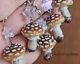 May include: A set of four mushroom charms featuring brown caps with white spots and white stems with dark brown bases. Each charm is attached to a silver-toned metal lobster clasp and a small, translucent, iridescent star-shaped bead.