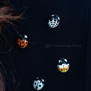 May include: Four small ladybug-shaped pins attached to a black fabric surface. The pins feature distinct designs: a white spider web, an orange jack-o'-lantern, a candy corn pattern, and a white ghost face. The text @KrintinCreations is visible.