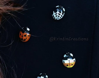 May include: Four small ladybug-shaped pins attached to a black fabric surface. The pins feature distinct designs: a white spider web, an orange jack-o'-lantern, a candy corn pattern, and a white ghost face. The text @KrintinCreations is visible.
