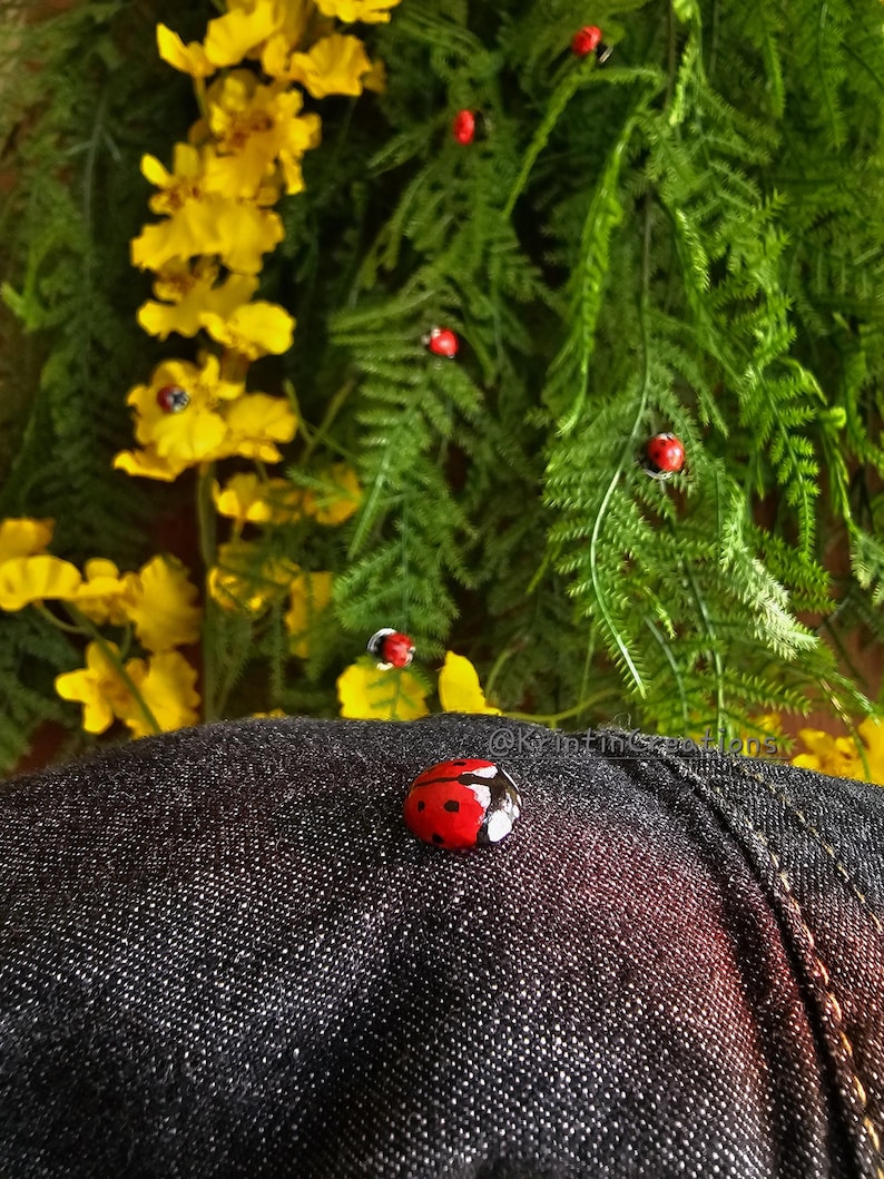 May include: A red ladybug with black spots is sitting on a dark blue denim fabric. The ladybug is in focus, while the denim fabric is out of focus. The background is a blurry image of yellow flowers and green leaves.