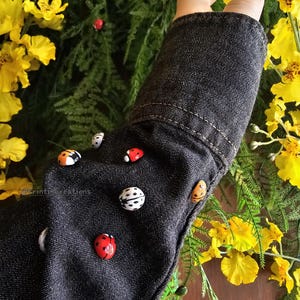 May include: A black denim jacket with red and black ladybug buttons. The jacket is surrounded by yellow flowers and green foliage.