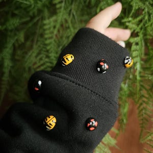 Puede incluir: Manga de sudadera negra adornada con varias cuentas redondas decorativas. Las cuentas presentan varios diseños en amarillo, negro y rojo. El fondo es una vista borrosa de follaje verde.