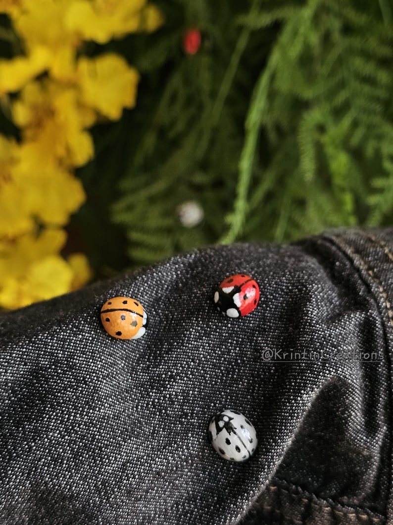 May include: Four ladybug pins on a dark denim jacket. The ladybugs are painted in different colors: red, orange, black, and white.