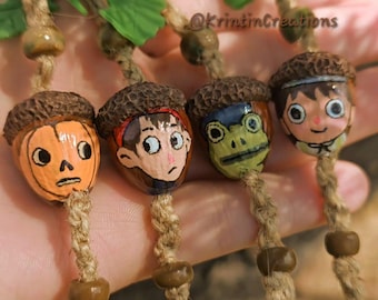 May include: Four hanging ornaments made from painted acorns with textured brown caps, each featuring a unique character face. The ornaments are suspended on braided jute twine with green plastic leaves and brown and green beads. Text reads @KrintinCreations.