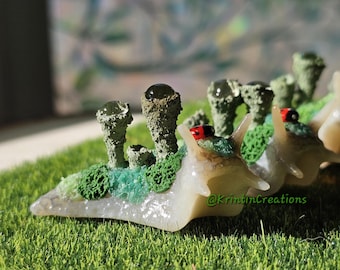 May include: A whimsical, handcrafted snail figurine with a translucent body and detailed green and brown mushroom-like structures on its back. The snail is set on a bed of artificial green grass, with the artist's name, @KritinCreations, visible.