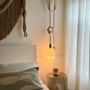 May include: A white rope light fixture hangs from a wooden wall mount. The light is on and illuminates a white bed with a grey patterned pillow. A wooden side table with a carved design sits next to the bed, with a small yellow ceramic cup on top.