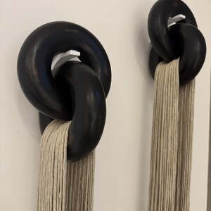 May include: Two wall-mounted displays featuring large, interlocking black circular rings suspended on white hooks. Each set of rings holds a long, vertical bundle of beige, multi-strand cord that hangs straight down from the bottom ring.