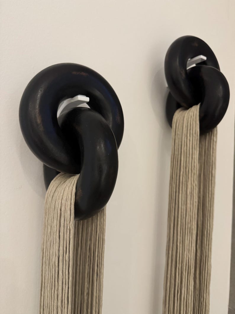 May include: Two wall-mounted displays featuring large, interlocking black circular rings suspended on white hooks. Each set of rings holds a long, vertical bundle of beige, multi-strand cord that hangs straight down from the bottom ring.