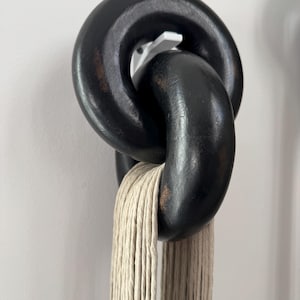 May include: Two interlocking, thick, matte black wooden chain links with visible brown wood grain distressing. The links are suspended from a white wall hook, and a bundle of light beige, multi-strand twisted cotton cord hangs through the bottom link.