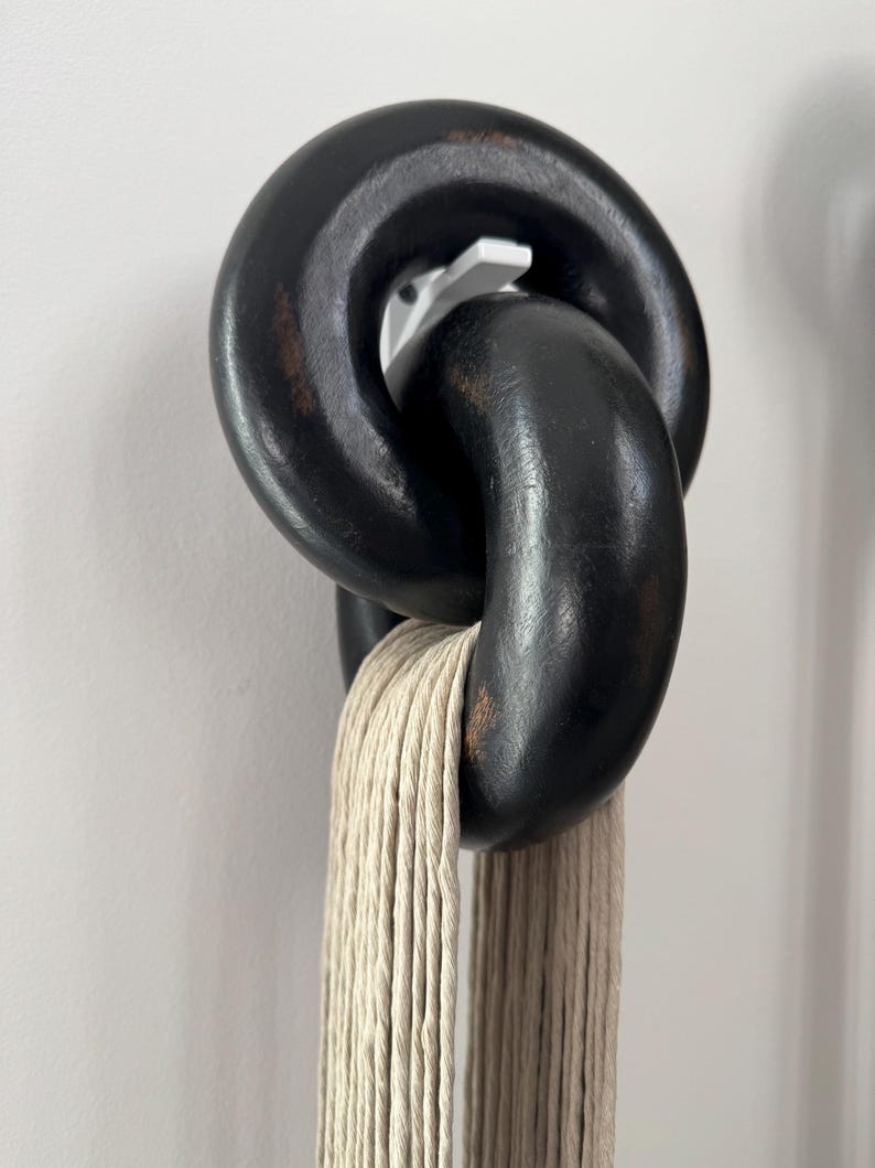 May include: Two interlocking, thick, matte black wooden chain links with visible brown wood grain distressing. The links are suspended from a white wall hook, and a bundle of light beige, multi-strand twisted cotton cord hangs through the bottom link.