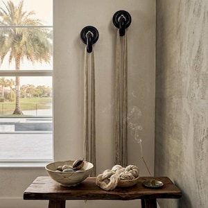 May include: Two black circular wall hooks mounted on a wall, each holding a long beige cord. Below, a wooden bench holds a ceramic bowl with stones, a small woven basket containing beige yarn bundles, and a metal incense holder with smoke.