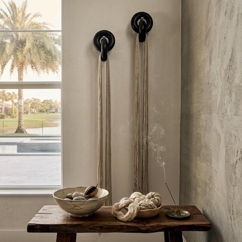 May include: Two black circular wall hooks mounted on a wall, each holding a long beige cord. Below, a wooden bench holds a ceramic bowl with stones, a small woven basket containing beige yarn bundles, and a metal incense holder with smoke.