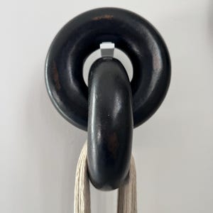 May include: A wall-mounted hook featuring two interlocking black circular rings with a distressed finish. The top ring is fixed to the wall, while the bottom ring hangs freely, supporting a length of light beige, multi-strand cotton cord.