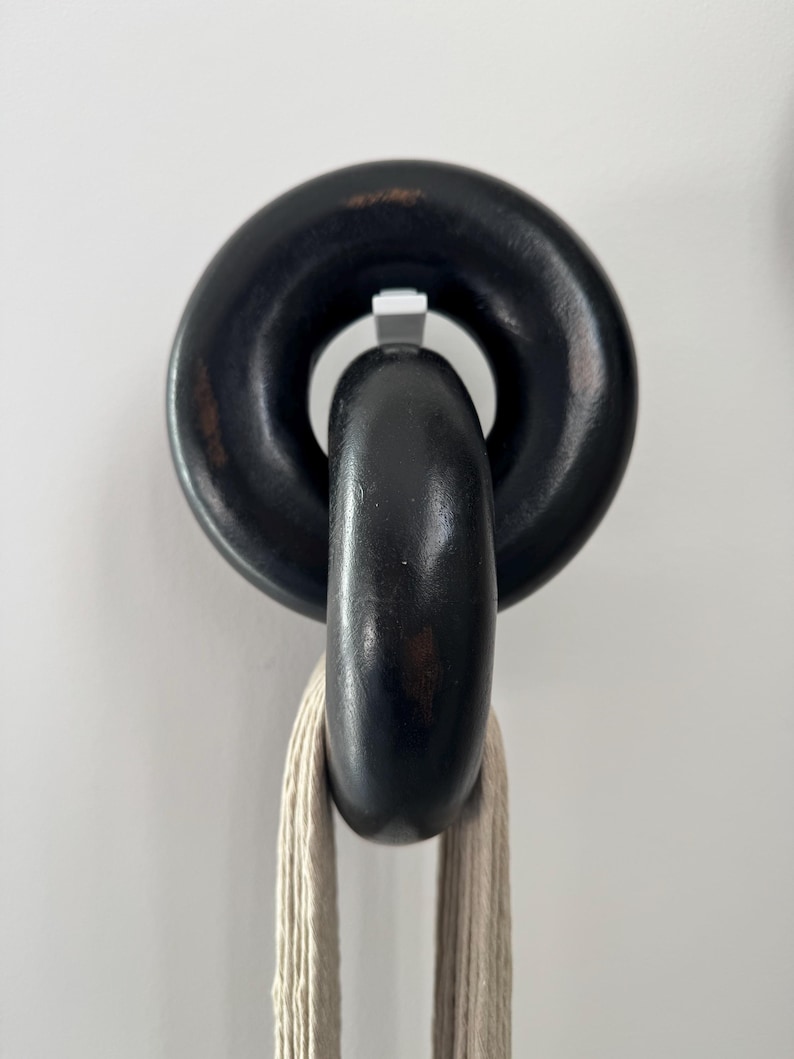 May include: A wall-mounted hook featuring two interlocking black circular rings with a distressed finish. The top ring is fixed to the wall, while the bottom ring hangs freely, supporting a length of light beige, multi-strand cotton cord.