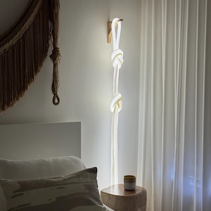 Puede incluir: Una lámpara de pared moderna y minimalista con un diseño único. La lámpara presenta una estructura similar a una cuerda blanca con dos grandes nudos, acentuados con dorado, colgando de un soporte de madera. La lámpara está iluminada, creando un ambiente cálido. Tema: decoración del hogar.