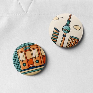 Könnte beinhalten: Zwei runde Pins mit Illustrationen von Berliner Sehenswürdigkeiten. Ein Pin zeigt einen roten Zug mit dem Wort "Pankow" an der Seite. Der andere Pin zeigt einen blauen und weißen Turm mit einem roten Dach und dem Text "Berlin" an der Seite.
