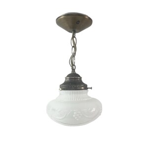 May include: Vintage pendant light with a white glass shade and bronze-colored metal accents. The shade has a decorative grape vine design. The light hangs from a chain and a ceiling mount.