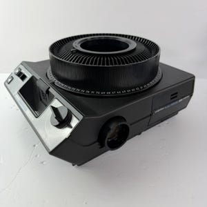 May include: A black Kodak Carousel 5500 slide projector featuring a circular tray on top with numbered slots, a front-facing lens, and a control panel with a black dial and switches. The device has a matte finish and a compact, rectangular design.