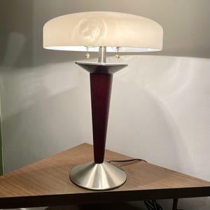 May include: A mid-century modern table lamp with a cream lampshade. The lamp has a dark red wooden base, a brushed metal stem, and a circular metal base. The lamp is plugged into an electrical outlet.