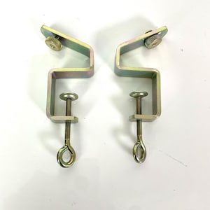 May include: Two metal C-clamps with a zinc-plated finish. Each clamp has a threaded rod with an eyelet, a screw for adjustment, and a right-angle design. The clamps are designed to secure objects.