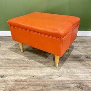 May include: An orange, square ottoman with four wooden legs. The legs have gold-colored metal tips. The ottoman is upholstered in a smooth, solid orange material. The design is mid-century modern.