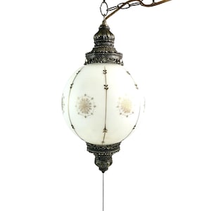 May include: A vintage, globe-shaped pendant light. The white glass globe has a floral design and is accented with dark metal filigree at the top and bottom. A pull chain hangs from the bottom. The fixture is suspended by a chain.