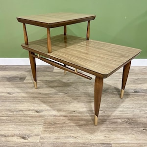 May include: A two-tiered side table in a mid-century modern style. The table features a rectangular top and a smaller, raised second tier. The legs are tapered with gold-coloured tips. The wood has a light brown, wood-grain pattern.