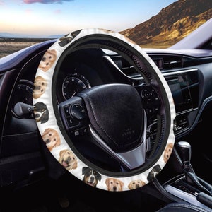 May include: White steering wheel cover with a black leather steering wheel. The cover is decorated with a pattern of various dog breeds, including golden retrievers, Bernese mountain dogs, and black and white dogs.